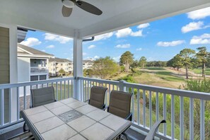 A covered balcony with a table and chairs, overlooking a golf course and residential buildings. - Beach at the Greens Retreat (Myrtle Beach)