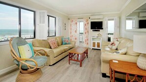 Ein Wohnzimmer mit Blick auf den Strand, ein Korbstuhl, ein beiges Sofa und ein Couchtisch aus Holz.