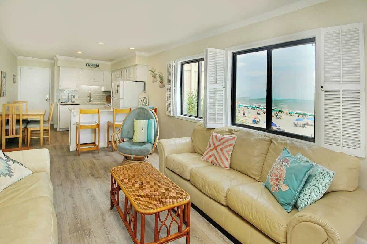 A living room with a sofa, a dining table, and chairs, a kitchen area, and a large window with beach view.
