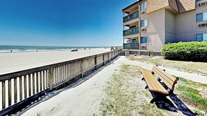 Ein Strandgrundstück mit Holzsteg, einer Bank und einem Gebäude mit Balkonen mit Blick auf den Ozean.