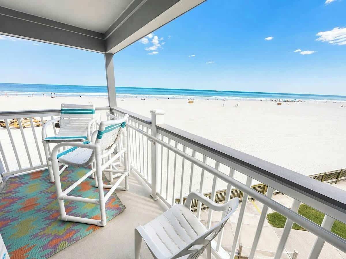 A balcony with white chairs and a colorful rug, overlooking a beach with clear blue water.