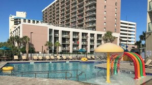 A swimming pool with a water feature, surrounded by lounge chairs and umbrellas, with a multi-story building in the background. - Sand Dunes North Tower #1510 (Myrtle Beach)