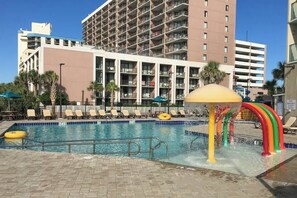 A swimming pool with a water feature, surrounded by lounge chairs and umbrellas, with a multi-story building in the background. - Sand Dunes North Tower #1510 (Myrtle Beach)
