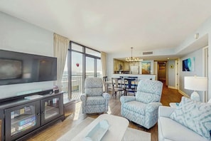 A modern living room with a flat-screen TV, two patterned armchairs, a white sofa, and a dining area with a chandelier. - Sand Dunes North Tower #1510 (Myrtle Beach)