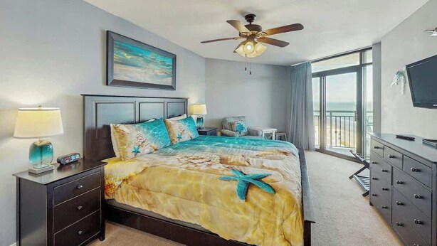 A hotel room with a bed, a ceiling fan, a TV, a nightstand, and a view of the ocean. - Sand Dunes North Tower #1510 (Myrtle Beach)