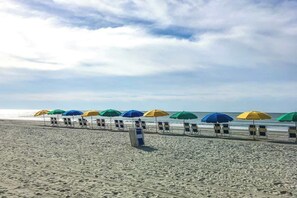 Ein Strand mit Reihen aus Liegestühlen und Sonnenschirmen, einem klaren Himmel und dem Meer im Hintergrund.