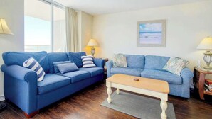 A living room with two blue sofas, a wooden coffee table, and a view of the sea through large windows. - North Hampton Oceanfront 1501 (Myrtle Beach)