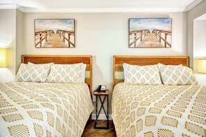 Ein Hotelzimmer mit zwei Betten, jeweils mit gemusterten Bettbezügen, einem kleinen Nachttisch mit Telefon und zwei an der Wand hängenden Bildern eines hölzernen Steg.