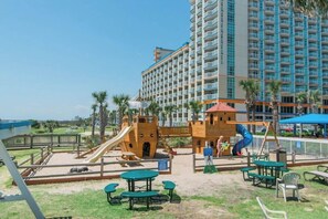 Een speelplaats met glijbanen en klimconstructies, omringd door picknicktafels en banken, met een hotel op de achtergrond.