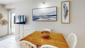 Un coin repas avec une table en bois, des chaises blanches, un téléviseur et des œuvres d’art encadrées accrochées au mur.