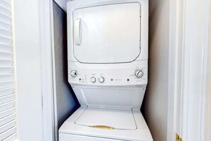 A compact, white, front-loading washing machine in a small laundry room with a door and a window. - Barefoot Sun & Golfing Fun (North Myrtle Beach)