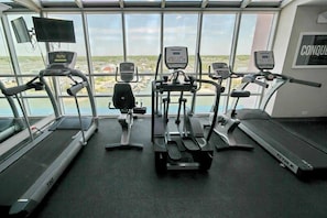 Ein Fitnessstudio mit Laufbändern, Crosstrainern und einem Hometrainer, mit Blick auf ein Gewässer und Gebäude durch große Fenster.