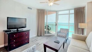 Ein Hotelzimmer mit Fernseher, einer dunklen Holzkombination, einer Couch, einem Couchtisch, einem Deckenventilator und einer Schiebetür zu einem Balkon mit Meerblick.