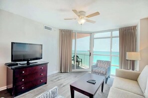 A hotel room with a television, a dark wooden dresser, a sofa, a coffee table, a ceiling fan, and a sliding glass door leading to a balcony with a view of the ocean. - Prince Resort Unit 1708 (North Myrtle Beach)