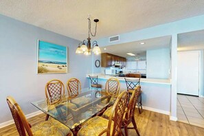 A dining area with a glass table and wooden chairs, a kitchen in the background, and a beach-themed painting on the wall. - Carolina Dunes Unit 603 (Myrtle Beach)