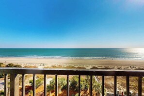 Blick auf den Strand vom Balkon mit Geländer, klarer Himmel und ruhiges Meer.
