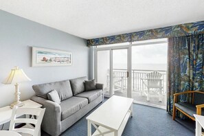 Ein Hotelzimmer mit grauem Sofa, einem weißen Tisch, einem Balkon mit weißen Stühlen und Meerblick.