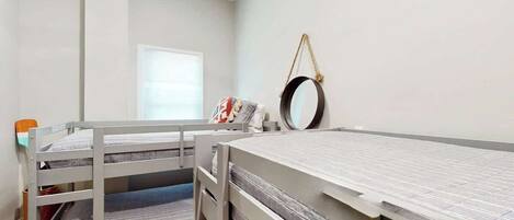 A room with bunk beds, a window with blinds, a round mirror on the wall, and a small table with a chair.