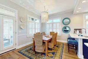 A dining area with a wooden table and woven chairs, a chandelier, and a patterned rug. - Coastal Escape at Litchfield by the Sea (Litchfield Beach)