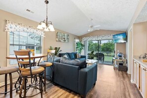A cozy living room with a dark sofa, wooden dining table, and chairs. There is a ceiling fan, a TV mounted on the wall, and a window with blinds. - Coastal Charm Unit 2G (Myrtle Beach)