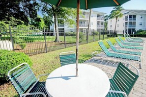 A patio area with a round table and chairs, a fenced garden, and a poolside lounge with chairs. - Coastal Charm Unit 2G (Myrtle Beach)