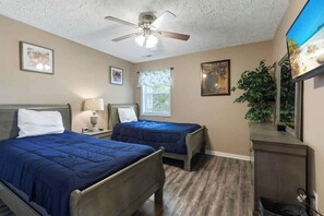Une chambre avec deux lits, un ventilateur de plafond, une fenêtre avec des stores et une plante.