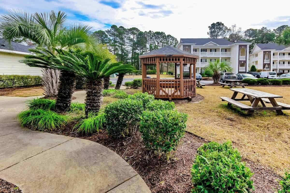 A gazebo in a landscaped outdoor area with a picnic table, surrounded by well-maintained shrubs and a palm tree.