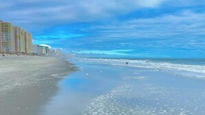 Ein Strand mit einer Reihe von Gebäuden links, einem klaren Himmel und ruhigen Wellen.