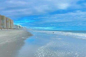 A beach with a row of buildings on the left, a clear sky, and calm sea waves. - Bay Watch Resort 0933 (North Myrtle Beach)