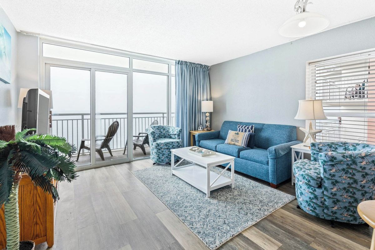 A living room with a blue sofa, matching armchairs, a coffee table, and a large window with curtains.