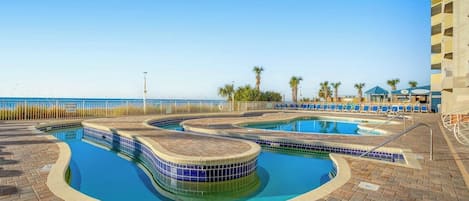 A pool area with multiple swimming pools, a beach view, and a building with balconies.