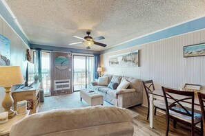Ein Wohnzimmer mit Deckenventilator, einer Couch, einem Esstisch mit Stühlen und Blick durch eine Glastür nach draußen.