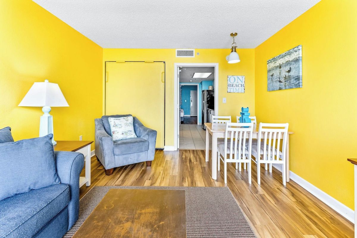 A room with yellow walls, a blue sofa, a wooden coffee table, a dining table with chairs, and a lamp.