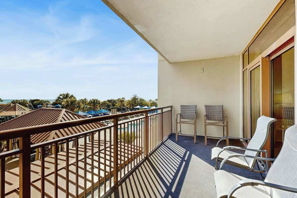 A balcony with a view of a pool and palm trees, two wicker chairs, and a glass door leading to an interior space.
