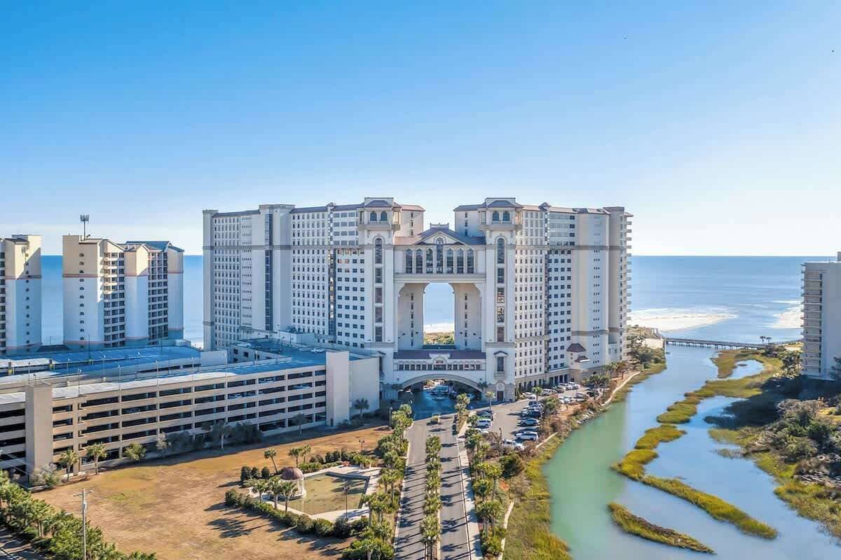 A high-rise hotel with a central archway, a parking garage, and a river running alongside.
