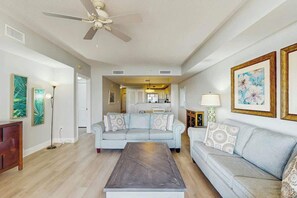 A living room with a ceiling fan, two sofas, a coffee table, and a kitchen in the background. - North Beach Towers 213 (North Myrtle Beach)