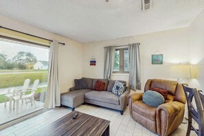 A living room with a sofa, armchair, and a table. There is a window with curtains, a sign on the wall, and a lamp. - Eagle Haven H1 Condo (Myrtle Beach)