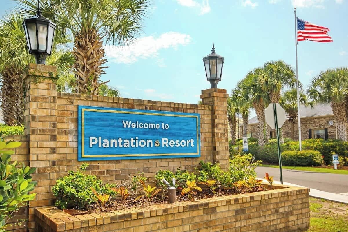 A sign welcoming to Plantation Resort with a brick wall, palm trees, and an American flag.