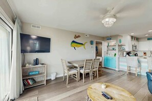 A modern living room with a flat-screen TV, a dining table with chairs, a wooden coffee table, and a kitchen area with a blue countertop. - Shore Breeze Unit 2 Condo (North Myrtle Beach)