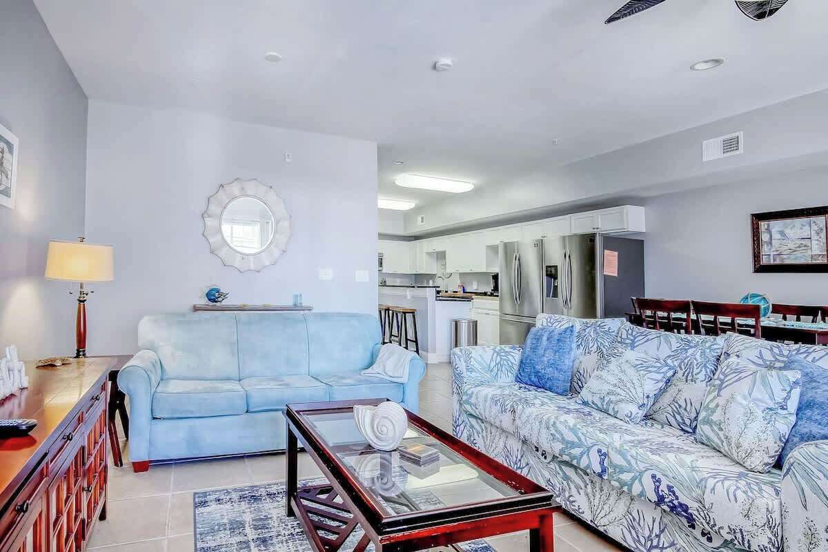 Una sala de estar con un sofá azul claro, un sofá estampado, una mesa de centro de vidrio y una cocina al fondo.