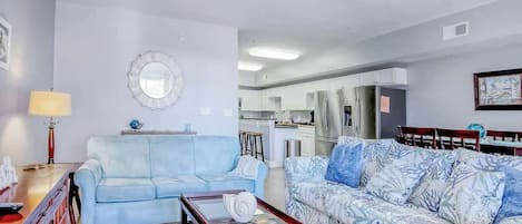 Una sala de estar con un sofá azul claro, un sofá estampado, una mesa de centro de vidrio y una cocina al fondo.