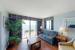 Ein Zimmer mit einer Couch, einem Tisch, einer Lampe, einer Zimmerpflanze und Blick auf das Meer durch große Fenster.