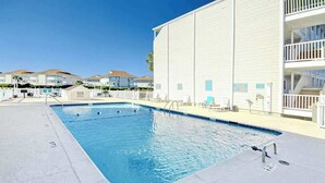 Ein Swimmingpool mit glasklarer, blauer Wasseroberfläche, umgeben von einem weißen Gebäude mit Balkonen, und einem wolkenlosen Himmel darüber.