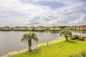 A hotel with a water body in front, palm trees, and a well-maintained lawn. - Beach Cottages at OD - 33B (North Myrtle Beach)