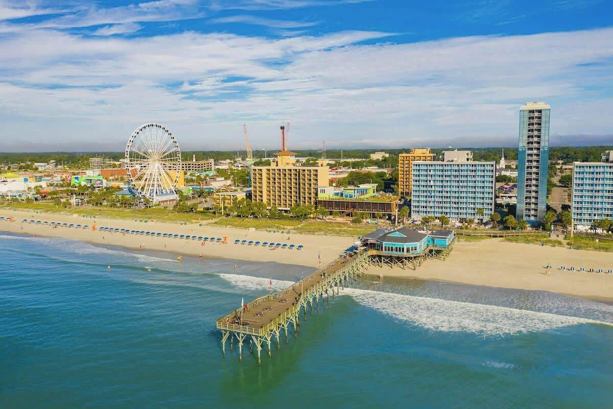 A coastal cityscape with a pier extending into the water, a large Ferris wheel, and modern buildings.