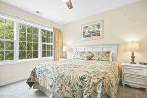 A bedroom with a large bed, a nightstand, a lamp, a window with curtains, and a painting on the wall. - The Havens Unit 1721 (North Myrtle Beach)