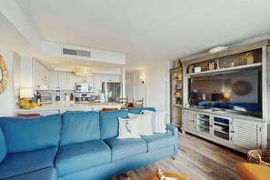A modern living room with a blue sofa, a television on a wooden cabinet, and a kitchen in the background. - Crescent Towers I - 102 (North Myrtle Beach)