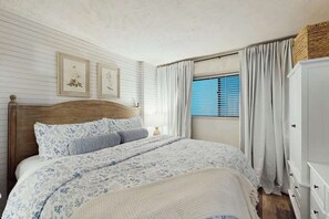 Ein Schlafzimmer mit einem hölzernen Kopfende, einem Bett mit floralem Bettzeug, einem Fenster mit blauen Fensterläden und einem weißen Kleiderschrank.