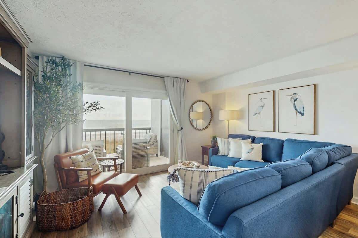 A living room with a blue sofa, a wooden chair, a table, and a large window with curtains.