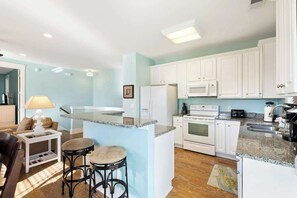 A kitchen with white cabinets, a granite countertop, a refrigerator, an oven, and a microwave. There is a bar stool and a lamp in the adjacent living area. - Madiera Magic Retreat (North Myrtle Beach)